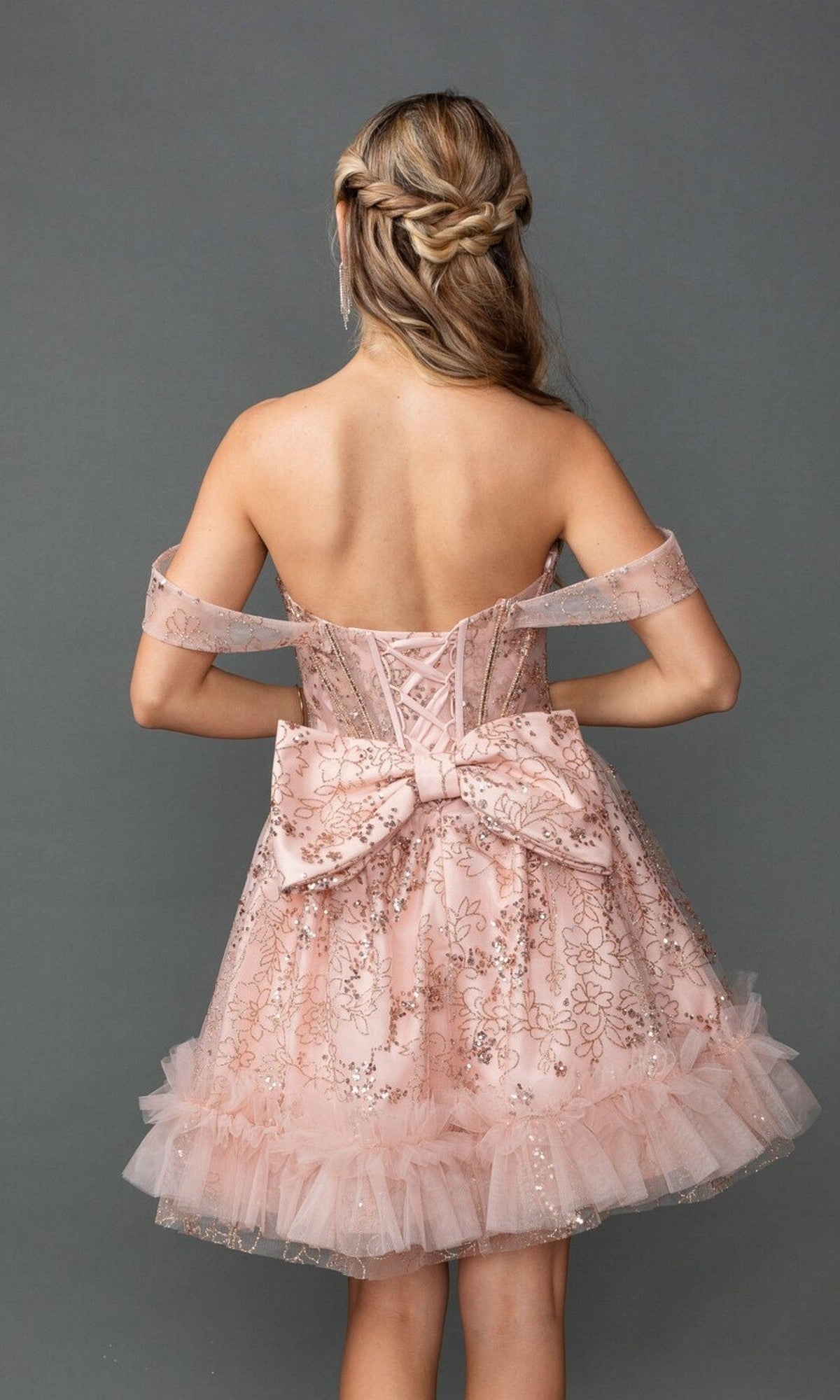 A woman with braided hair models the Dancing Queen 3422 short prom dress—a pink, off-the-shoulder style with a corset bodice, sequin floral print, and a large bow—photographed from the back against a gray backdrop.